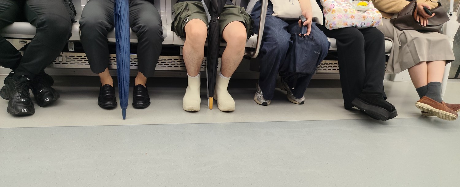 Füße nebeneinander auf einer Sitzbank in der Seouler U-Bahn: schwarze Sneaker, Loafer, weiße Gummi-Regenstiefel und weitere Sneaker, dazu angelehnte Regenschirme