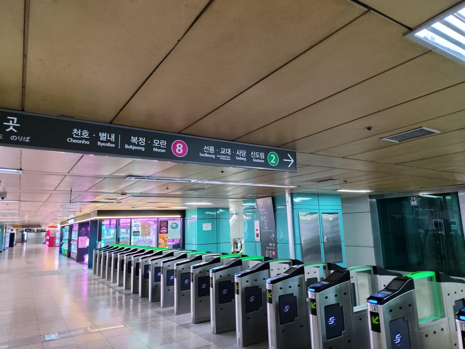 Lange Reihe von U-Bahn-Sperren an einem Umsteigebahnhof in Seoul, darüber bunte Schilder für Linie 8 (pink) und Linie 2 (grün), seitlich leuchtende Läden